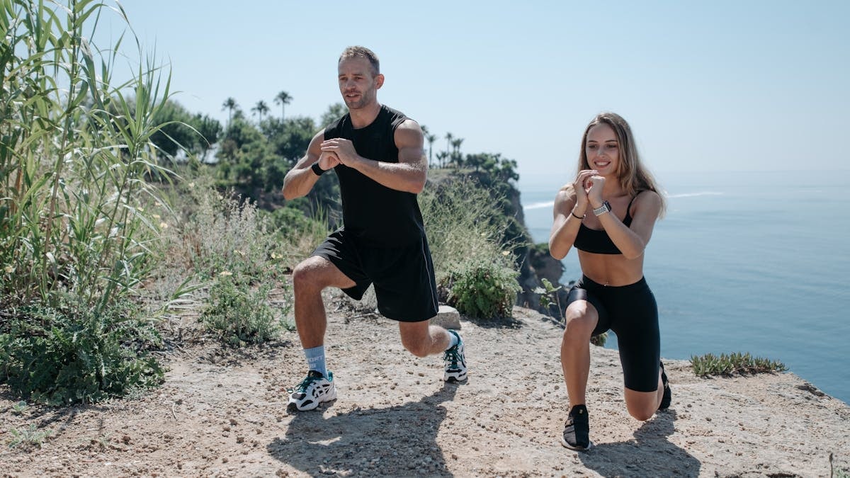 A man and woman exercising on a rocky cliff overlooking the ocean on a sunny day.